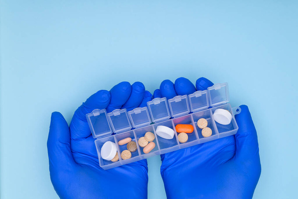 Pill box with medicines in the hands of the doctor.Hands of the doctor in blue gloves, hold in his hands box with medicines for every day, on blue background. Use of pills as prescribed by doctor. - Photo, image
