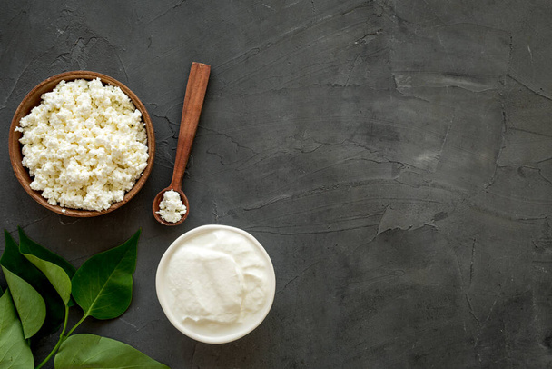 Cottage cheese in wooden bowl with green summer branch on dark kitchen table. Top view  - Фото, изображение