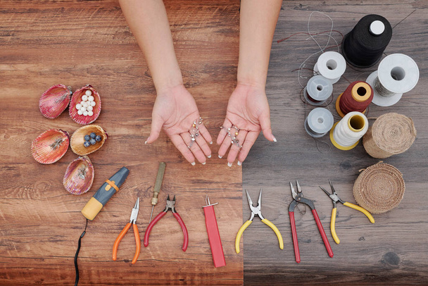 Manos de mujer mostrando nuevos pendientes que hizo con herramientas, hilos y perlas alrededor, vista desde arriba
 - Foto, imagen