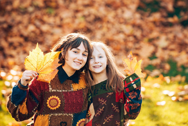 Two sisters girls with autumn orange leaves. Healthy and natural woman beauty. Smiling young girl with sister with an orange leaf in hands on autumn foliage background in the forest. Sisters portrait. - Photo, image