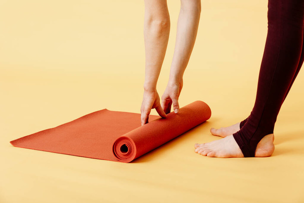 Gros plan de la jeune femme attrayante pliant yoga rouge ou tapis de remise en forme après avoir travaillé sur fond jaune. Yoga et mode de vie sain concept. Plan horizontal. Plan studio - Photo, image