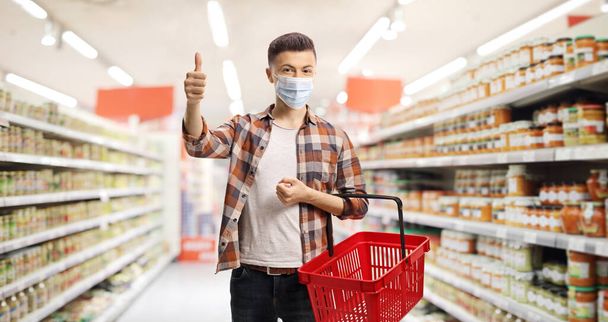 Guy in a supermarket with a protective face mask and a shopping basket showing thumbs up - Zdjęcie, obraz