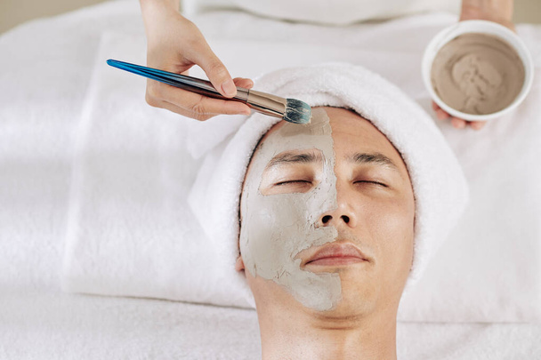 Kozmetik uzmanı erkek müşterinin yüzüne kilden maske takıyor. - Fotoğraf, Görsel