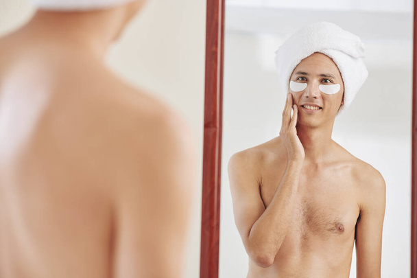 Feliz hombre sin camisa con toalla en la cabeza y bajo parches de ojos tocando la cara y mirando el espejo del baño
 - Foto, imagen