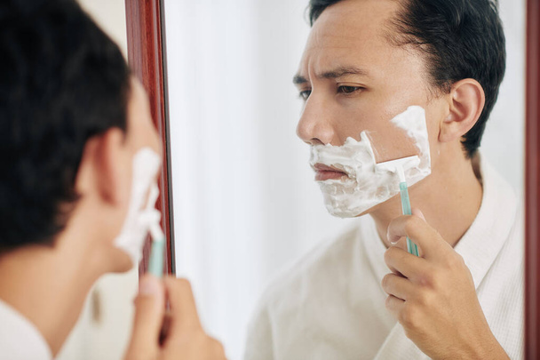 Serious frowning man looking at mirror when shaving in the morning - Photo, image