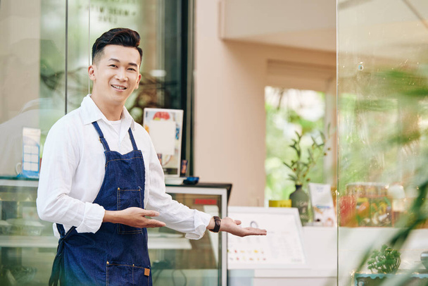 Porträt eines jungen positiven Vietnamesen, der Kunden in seinem Café begrüßt - Foto, Bild