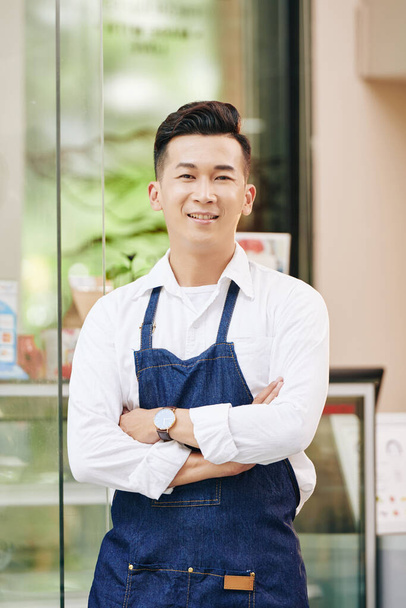 Retrato de garçom sorrindo positivo em denim avental de pé na frente do café com os braços cruzados
 - Foto, Imagem