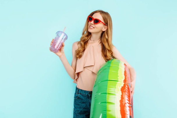 happy beautiful girl dressed in a swimsuit and sunglasses holds a cocktail and an inflatable swimming circle isolated on a blue background - 写真・画像
