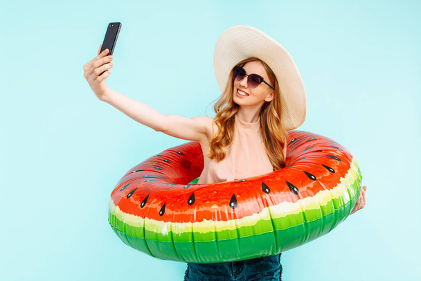 attractive smiling girl in a swimsuit and a summer hat holding an inflatable swimming circle and taking a selfie on a mobile phone on an blue background - Fotó, kép