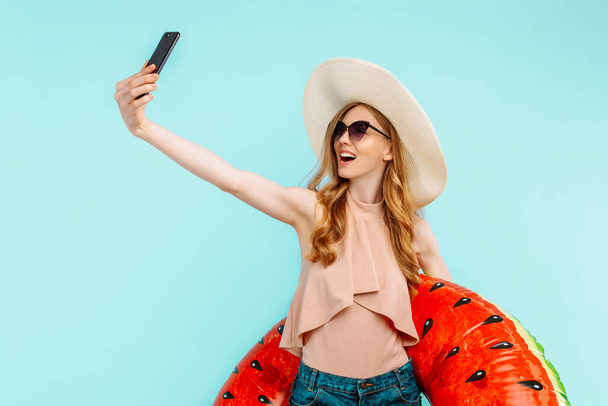 attractive smiling girl in a swimsuit and a summer hat holding an inflatable swimming circle and taking a selfie on a mobile phone on an isolated blue background - Photo, Image