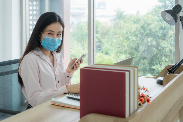 美しいアジアの女性は、マスクを着用し、自宅から動作するようにラップトップを使用しています。社会的距離は、コロナウイルスまたはCOVID-19の普及による新しい正常なものです。. - 写真・画像