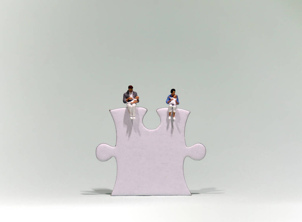 Un hombre en miniatura y una mujer en miniatura sentados encima de una pieza de rompecabezas sosteniendo a un bebé
.  - Foto, Imagen