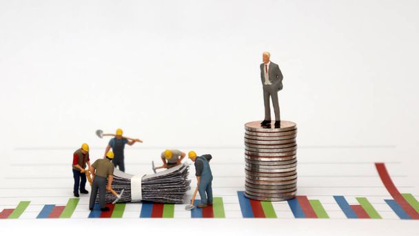 A man dressed in a suit standing on top of a pile of coins against a graph and miniature workers at a construction site below it. - Photo, Image