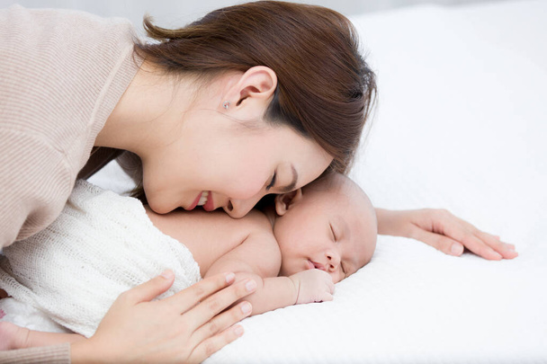 Piękna mama kłamie i delikatnie przytula noworodka delikatnie, podczas gdy niemowlę śpi na białym łóżku. Azji matka całuje i dotyka na plecach dzieci z miłości i opieki. - Zdjęcie, obraz