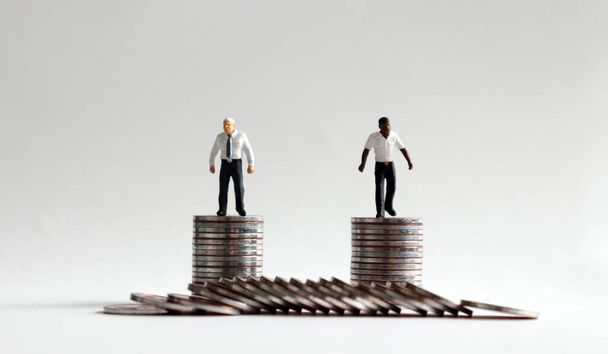 Un hombre blanco en miniatura y un hombre negro en miniatura de pie sobre una pila de monedas de la misma altura
. - Foto, imagen