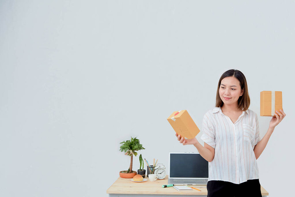 Азиатка держит коробку с посылками, работает в домашнем офисе. Онлайн-маркетинг, стартап МСП предприниматель или внештатная концепция
 - Фото, изображение