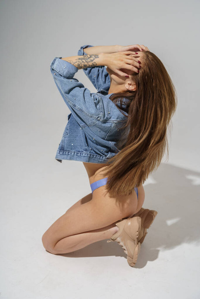 Attractive long-haired brunette in blue lingerie and blue jeans jacket posing on gray background   - 写真・画像