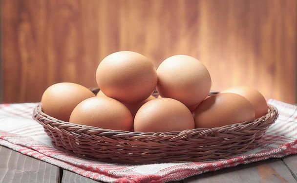 Chicken eggs in a wicker basket on an old wooden table with a blurred background. - Фото, изображение
