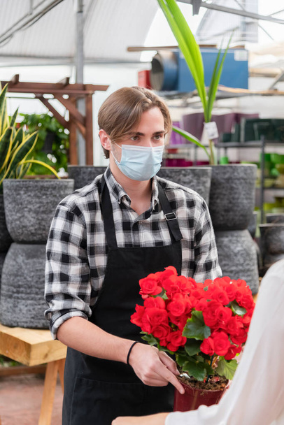 Young handsome clerk wearing an apron and a surgical mask holding a plant on an out of focus background. Garden center and shopping concept. - Photo, Image