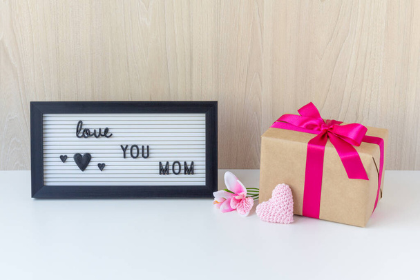 Διακοσμητική πινακίδα με το μήνυμα: Σ 'αγαπώ μαμά. Δίπλα έχουν ένα λουλούδι, μια καρδιά βελονάκι και ένα όμορφο δώρο τυλιγμένο σε μια ροζ κορδέλα. Ξύλινο φόντο και λευκή βάση. - Φωτογραφία, εικόνα