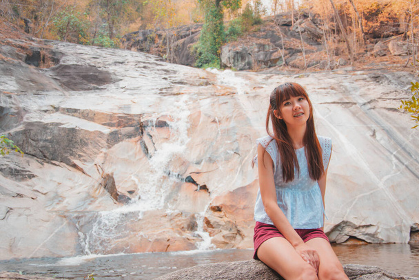 Mulher asiática viajantes estão sorrindo e relaxa sentado na pedra em frente à cachoeira em Namtok Kaew Cha
 - Foto, Imagem