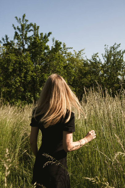 Κορίτσι με μαύρο φόρεμα περπατάει στη φύση. Ξανθιά σε κίνηση στο πεδίο. Ένα νεαρό κορίτσι απολαμβάνει το καλοκαίρι. - Φωτογραφία, εικόνα