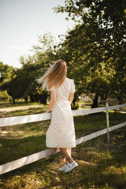 Girl in a white dress walking in nature. Blonde in motion on a farm. A young girl enjoy the summer. - Foto, afbeelding