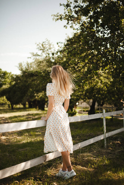 自然の中を歩く白いドレスの少女。農場でブロンドが動いてる。若い女の子は夏を楽しむ. - 写真・画像