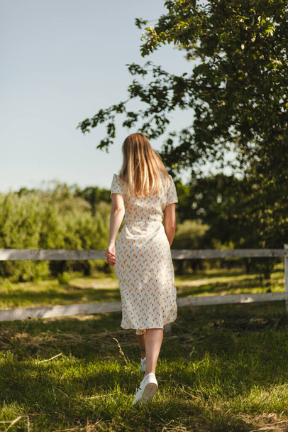 Egy fehér ruhás lány sétál a természetben. Szőke mozgásban egy farmon. Egy fiatal lány élvezi a nyarat.. - Fotó, kép