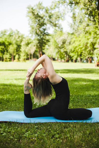 Portret van een prachtige jonge vrouw die buiten yoga beoefent in een park. Mooi meisje in een zwart mager pak. Kalmeer en ontspan, vrouwelijk geluk. Horizontale, wazige achtergrond. - Foto, afbeelding