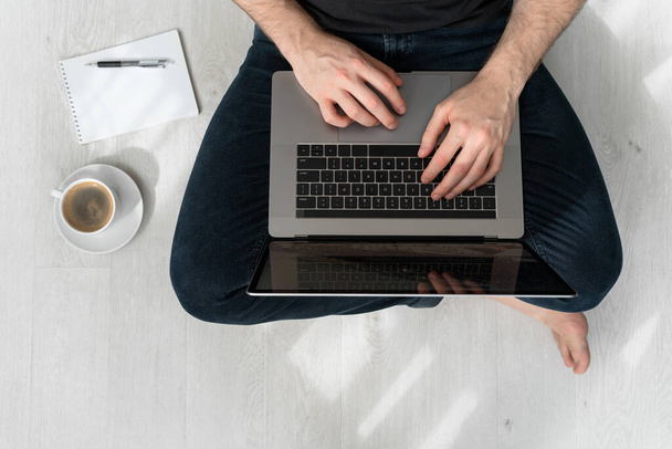 Man sitting on floor with laptop, notebook, pen and cup of coffee. Online office. Remote work. - Photo, image
