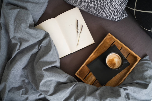 Black cup of coffee on notebook on wooden tray and open book with lavender flowers on bed with grey blanket and black and white pillows. Morning ritual. Breakfast in bed. - Photo, Image