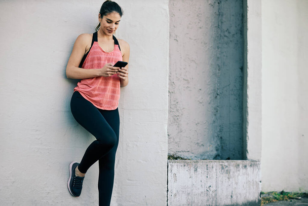 Genç ve yakışıklı sporcu kadın akıllı telefon kullanarak koşmaya ve duvara yaslanmaya ara veriyor.. - Fotoğraf, Görsel