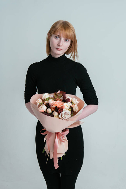 Portrait of young female florist with bouquet of flowers in peach foamiran package in the hands on a light background. Rosa, cotinus, brunia, lagurus. - Фото, изображение