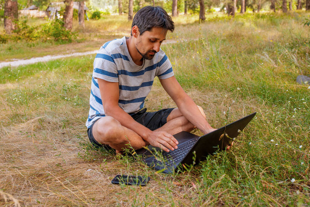 A young man with a beard sits on the grass and works on the computer. Concept - freelance outdoors in pleasure - Фото, зображення