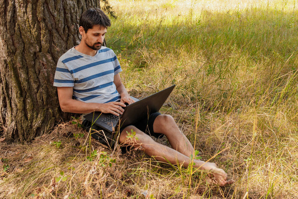 A young man with a beard sits on the grass and works on the computer. Concept - freelance outdoors in pleasure - 写真・画像