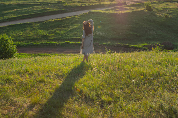 On the field is a barefoot girl in a gray dress at sunset. Back view - Zdjęcie, obraz