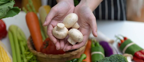 Закройте вид на руки, держащие белые грибы шампиньон, стоя за столом с другими свежими фермерскими овощами
 - Фото, изображение