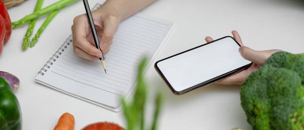 Cropped shot of female noting diet plan while looking information on mock-up smartphone on kitchen table with fresh vegetables  - Foto, afbeelding