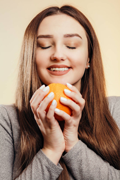 portrait of a beautiful positive girl with long hair and orange, a young woman smiling and holding fruit near face on studio background, healthy eating concept - Foto, afbeelding