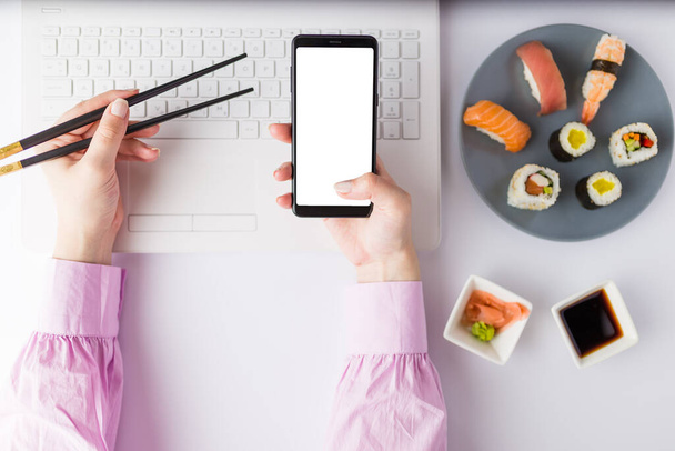 Flat lay of asian sushi set with laptop and female hand holding chopsticks and blank screen mobile phone. Healthy meal at work food delivery concept. - Zdjęcie, obraz