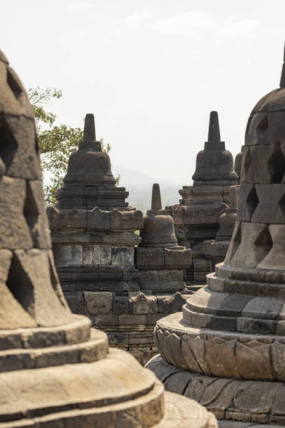 Orta Java, Endonezya 'daki Borobudur tapınağının farklı seviyelerindeki denge ve duvarlarına taç takan çeşitli stupaların ayrıntıları, arka planda, çevredeki manzaranın parçacıkları.. - Fotoğraf, Görsel