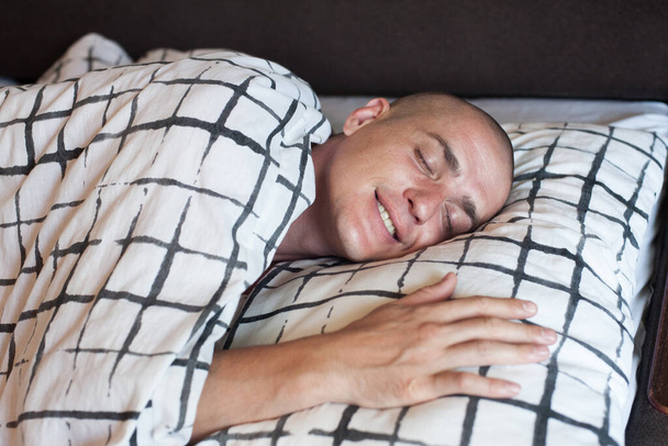Ένας άντρας με ευρωπαϊκή εμφάνιση κοιμάται και χαμογελά στον ύπνο του με ελαφριά σεντόνια. - Φωτογραφία, εικόνα