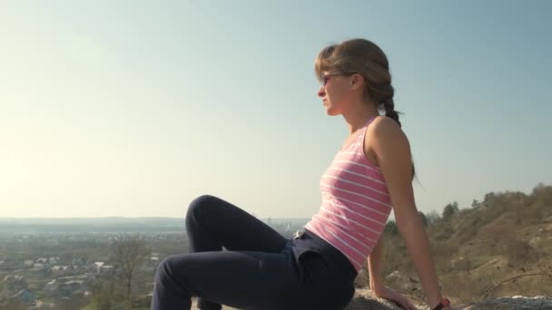 Mladá uvolněná žena sedí venku na velkém kameni a užívá si teplého letního dne. Dívka meditující a relaxující na přírodě při západu slunce. - Záběry, video