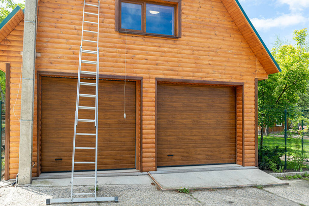Новый современный гараж для двух автомобилей строительной площадки окончательный разрез с деревянной стеной сайдинга. Высокое профессиональное алюминиевое лестничное оборудование на фасаде здания
 - Фото, изображение
