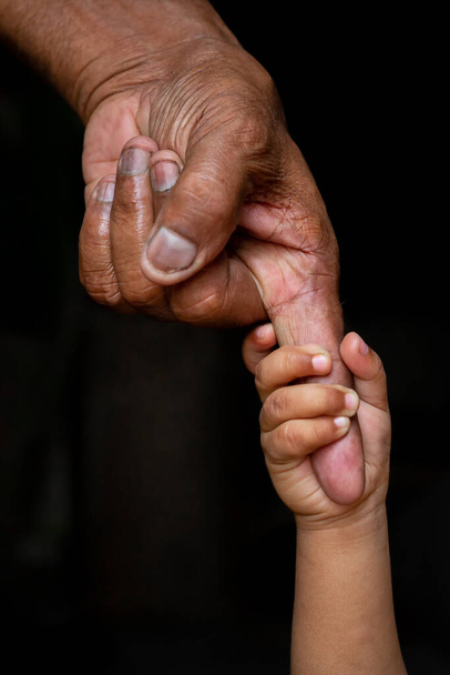 Las manos de un bebé agarrándose con fuerza al dedo mayor de un anciano. Familia, Generación, Apoyo y concepto de personas. Fondo oscuro
. - Foto, Imagen