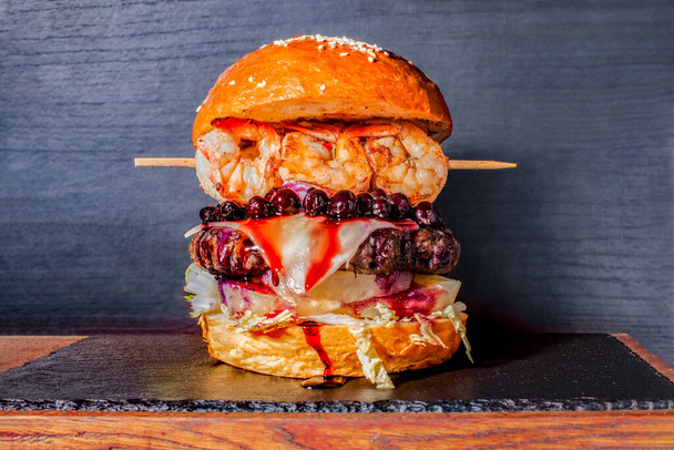 Unusual Huge burger with beef, shrimp, pineapple, blackcurrant, cheese, fresh vegetables and sauces on plate over wooden background. Craft beef big burger on a board with a stone and wooden deck - Zdjęcie, obraz