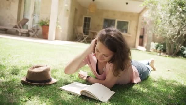 Young happy woman lying on a lawn in country house and makes notes in a notebook - Materiał filmowy, wideo