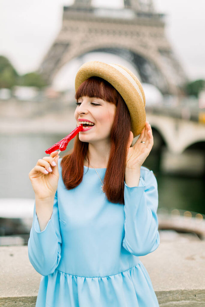 Young charming red haired laughing woman, wearing straw hat and elegant blue dress, posing for photo, biting red lollipop, while standing on Eiffel tower background and river Seine - Foto, immagini