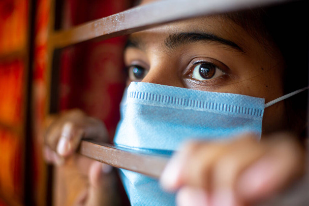 Ένα νεαρό κορίτσι από την Ασία που βαριέται και φοράει μάσκα προστασίας κοιτάζοντας μέσα από το παράθυρο να είναι σε καραντίνα στο σπίτι κατά τη διάρκεια της επιδημίας του κορωνοϊού. Κοντινές προβολές. - Φωτογραφία, εικόνα